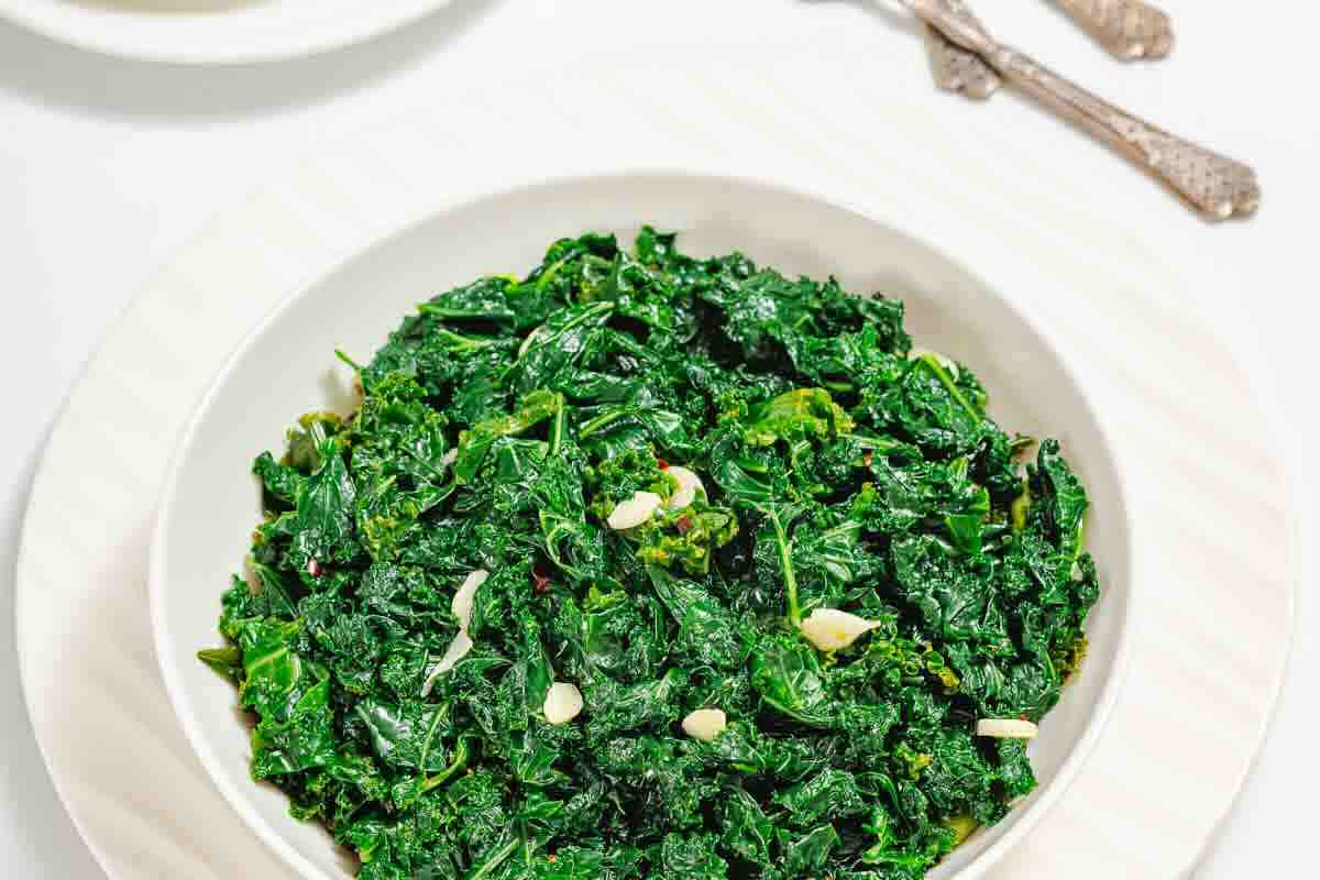 A white bowl filled with sauteed kale and slices of garlic, placed on a white plate with two silver forks in the background.
