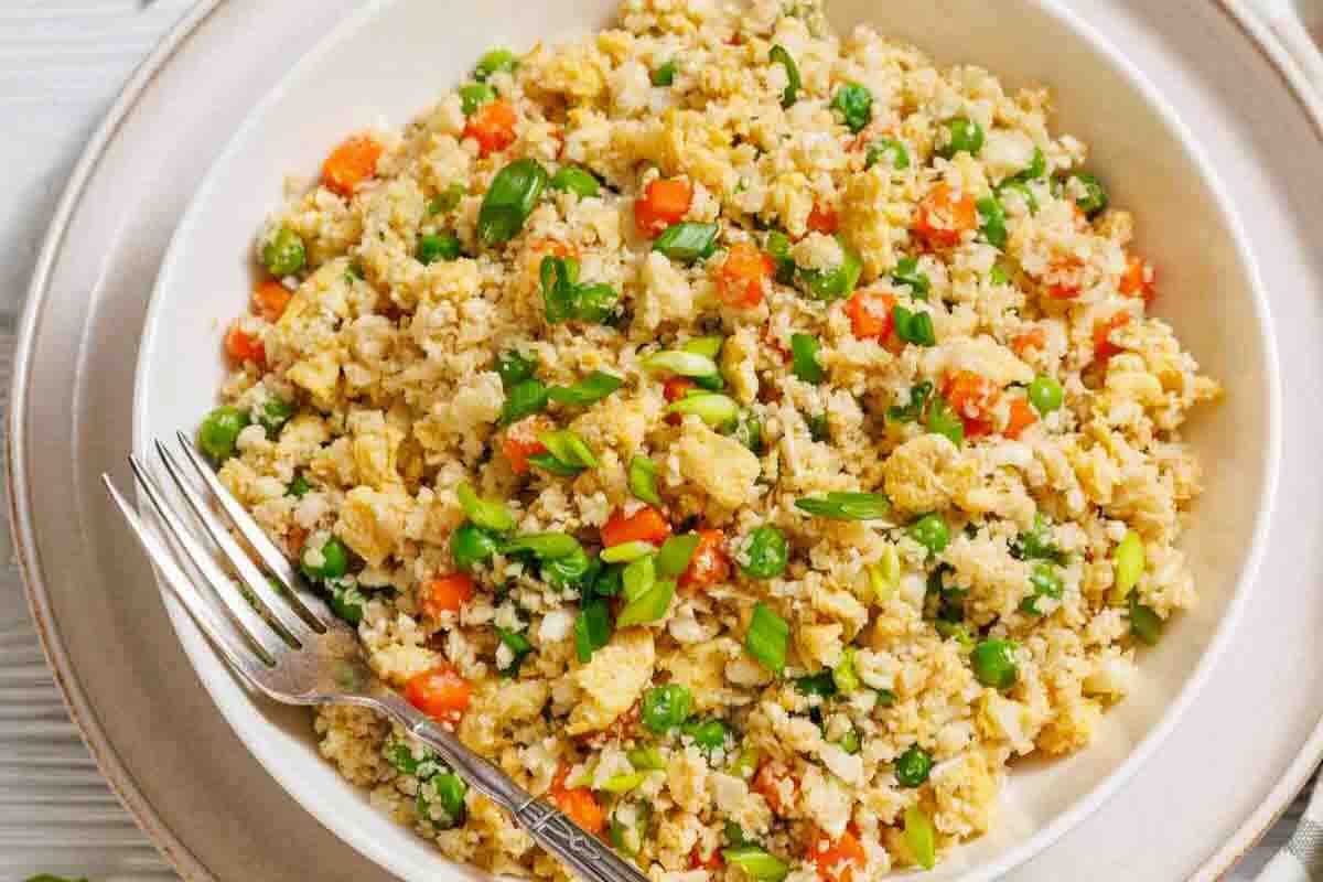 A bowl of cauliflower fried rice with peas, carrots, scrambled eggs, and chopped green onions, served with a fork.