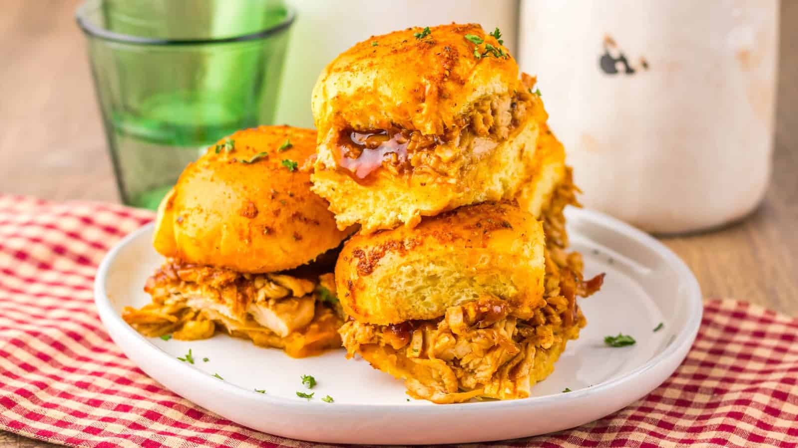 Three BBQ chicken sliders stacked on a white plate, with a red checked cloth and a green glass nearby.