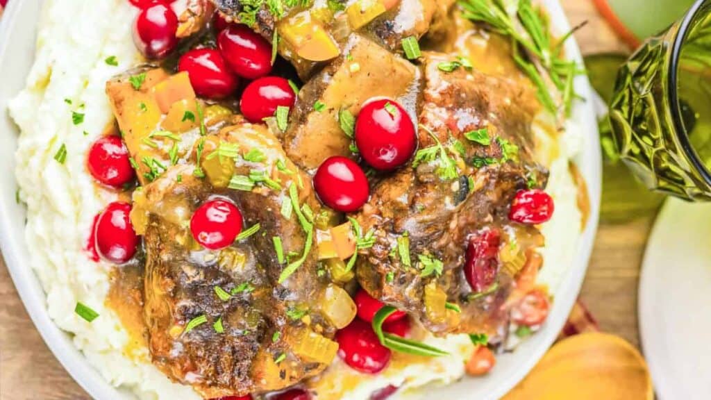 A plate of mashed potatoes topped with braised meat, cranberries, chopped herbs, and gravy.