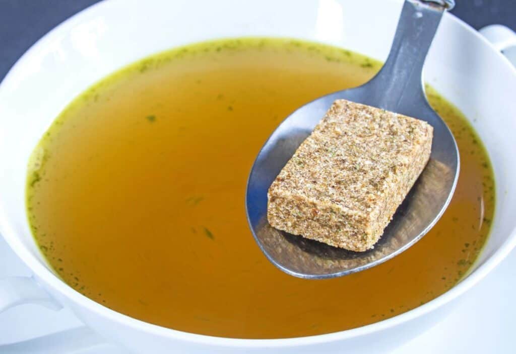 A metal spoon holding a brown bouillon cube is positioned over a bowl of clear broth.