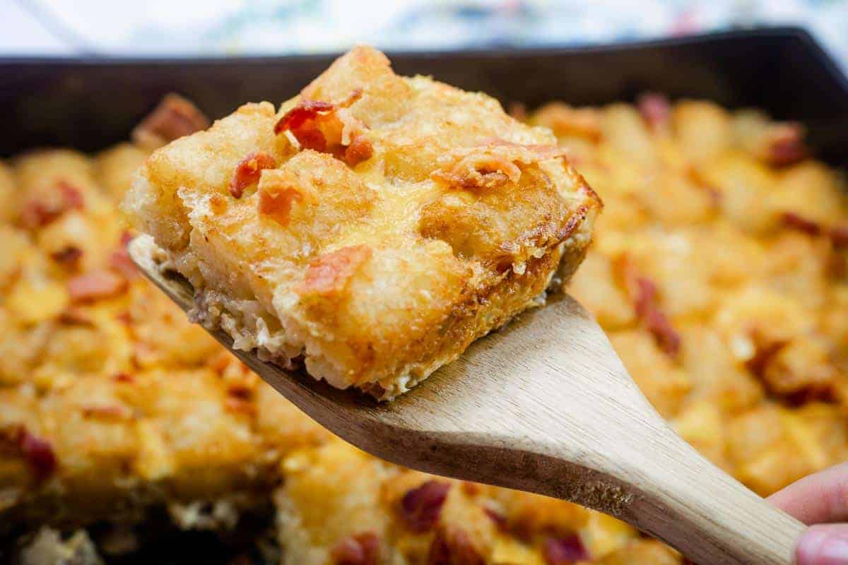 A wooden spatula holds up a square piece of Tater Tot Breakfast Casserole topped with crispy bits.