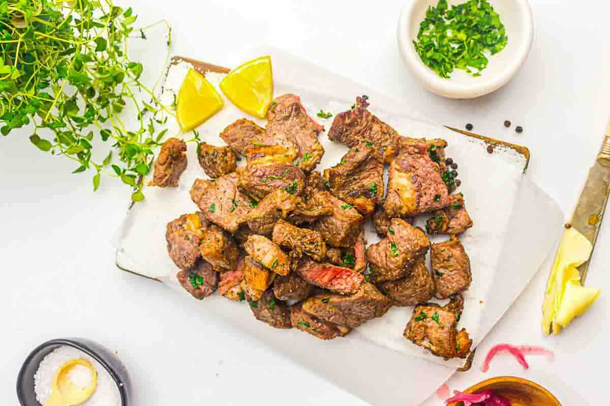 Grilled steak bites garnished with herbs on a white cutting board, served with lemon wedges and surrounded by small bowls of seasoning and chopped herbs.