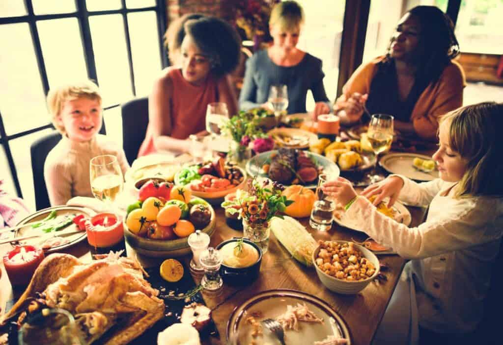 A group of people, including children and adults, sit around a table enjoying a meal together, sharing dishes and drinks that foster connection and warmth.