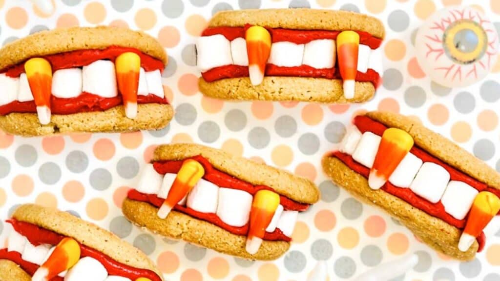 Five Halloween-themed cookie sandwiches are decorated with marshmallows and candy corn to resemble mouths with teeth and fangs, placed on a polka dot background.