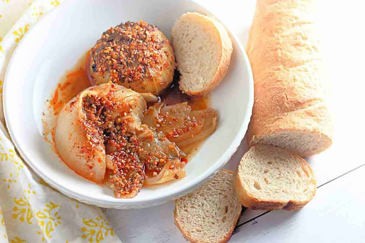 A bowl of cooked onions topped with spices and oil, served with sliced baguette on a white surface.