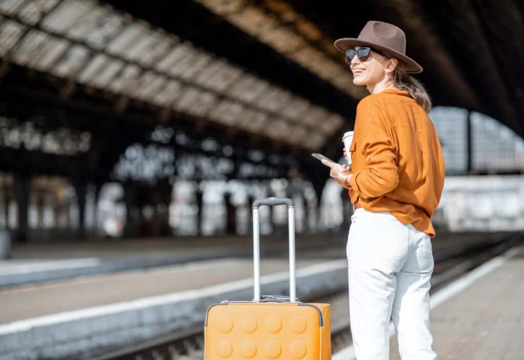Woman in a brown hat and sunglasses stands on a train platform with a yellow suitcase, coffee cup, and phone&mdash;ready to discover detour destinations, she faces away from the tracks with adventure in mind.
