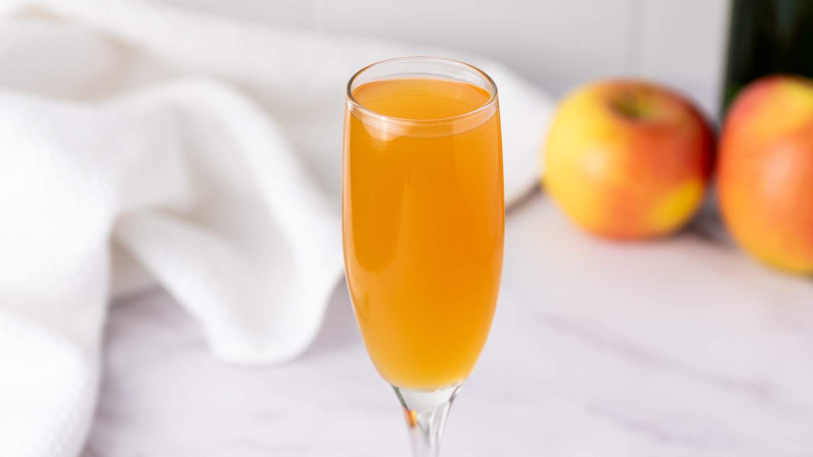 A champagne flute filled with orange-colored mimosa sits on a marble surface, with two apples and a white cloth in the background.