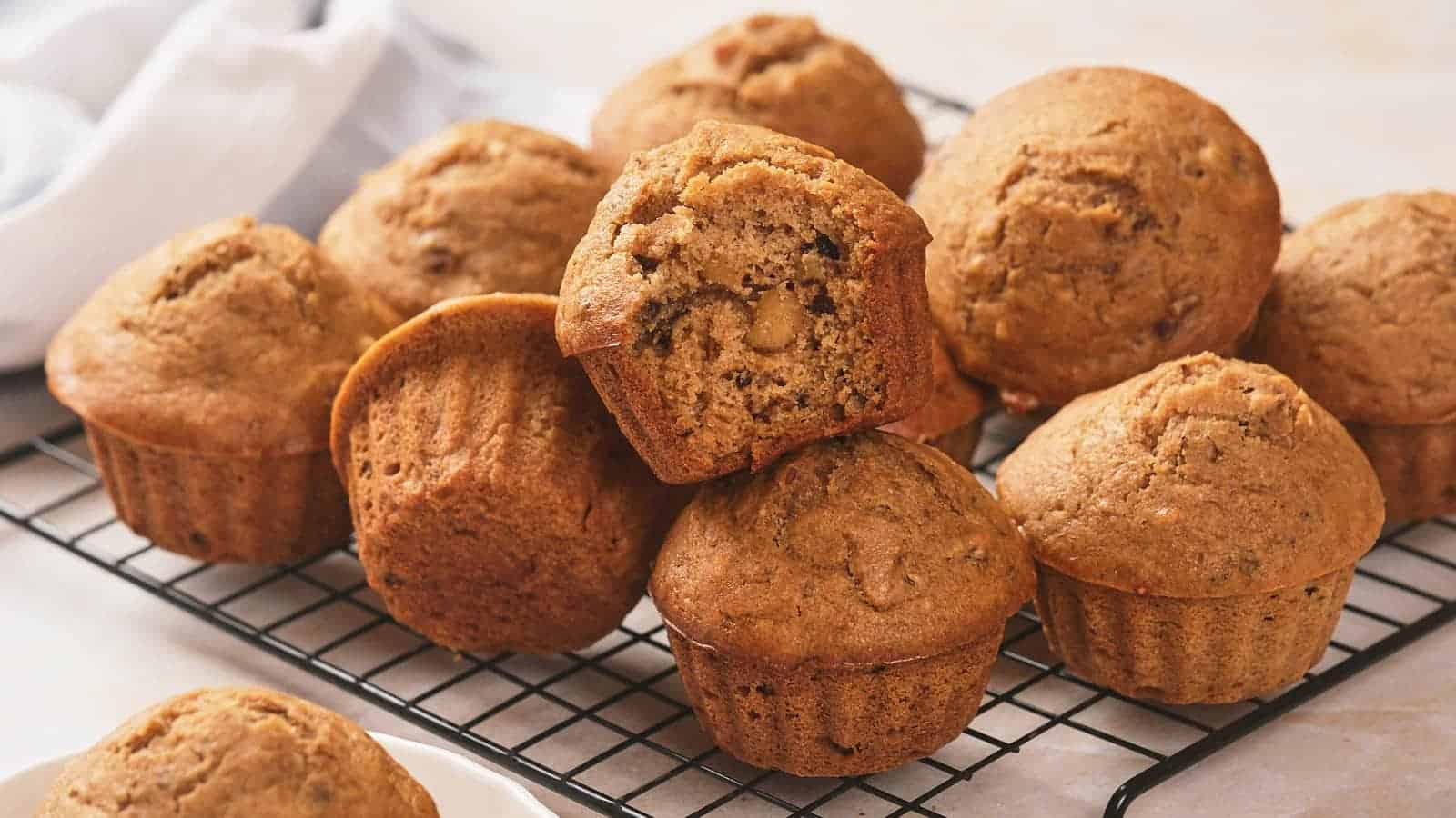 A batch of brown muffins rests on a cooling rack, with one muffin on top showing its crumb and nut pieces inside.