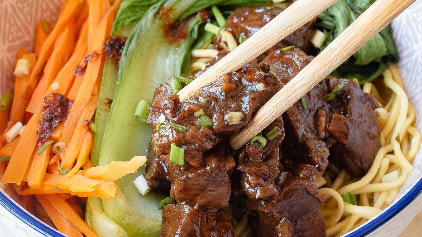A close-up of chopsticks holding pieces of braised beef over noodles, garnished with sliced carrots, green onions, and bok choy in a bowl.