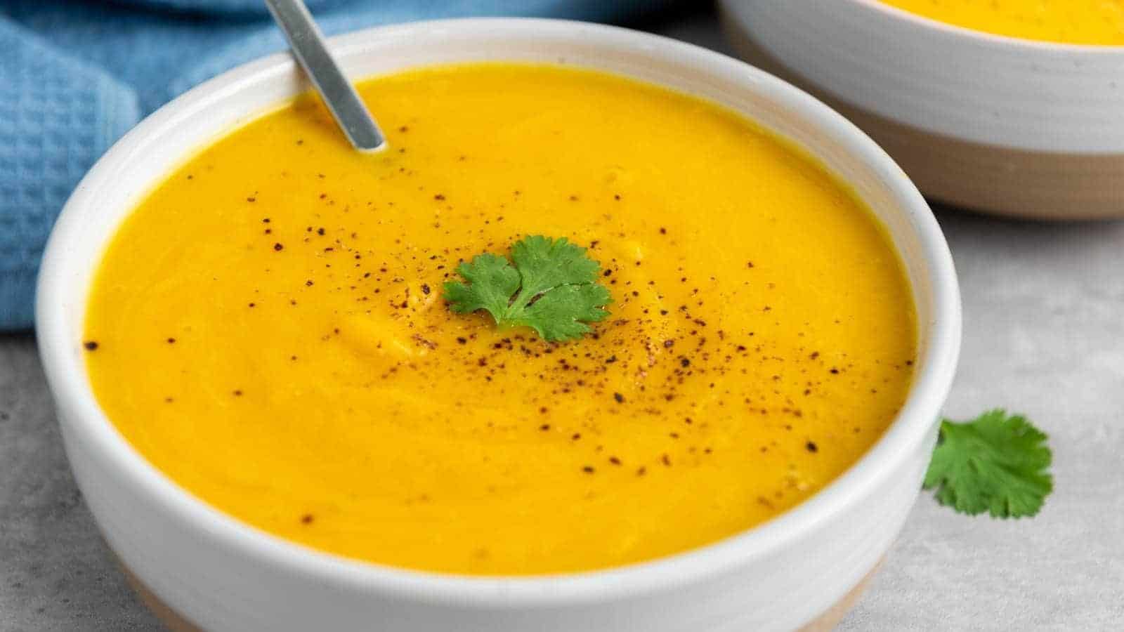 A bowl of smooth butternut squash soup, garnished with black pepper and a sprig of cilantro, with a spoon resting inside the bowl.