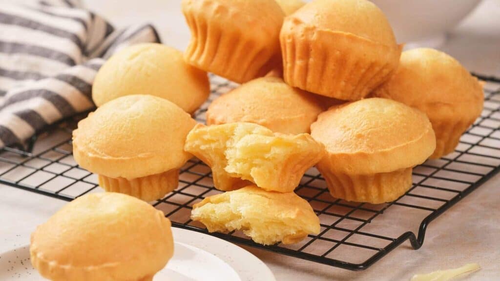Several golden cheese bread muffins are stacked on a cooling rack, with one muffin broken open to show its soft, airy interior. A striped cloth is visible in the background.