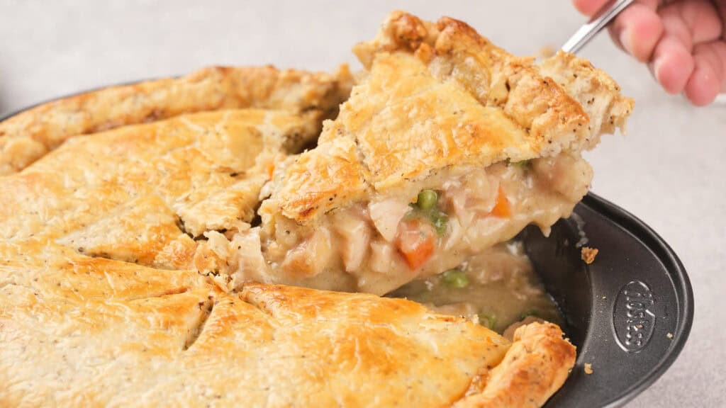 A hand lifts a slice of chicken pot pie from a pan, revealing a creamy filling with chicken, carrots, peas, and a golden, flaky crust.