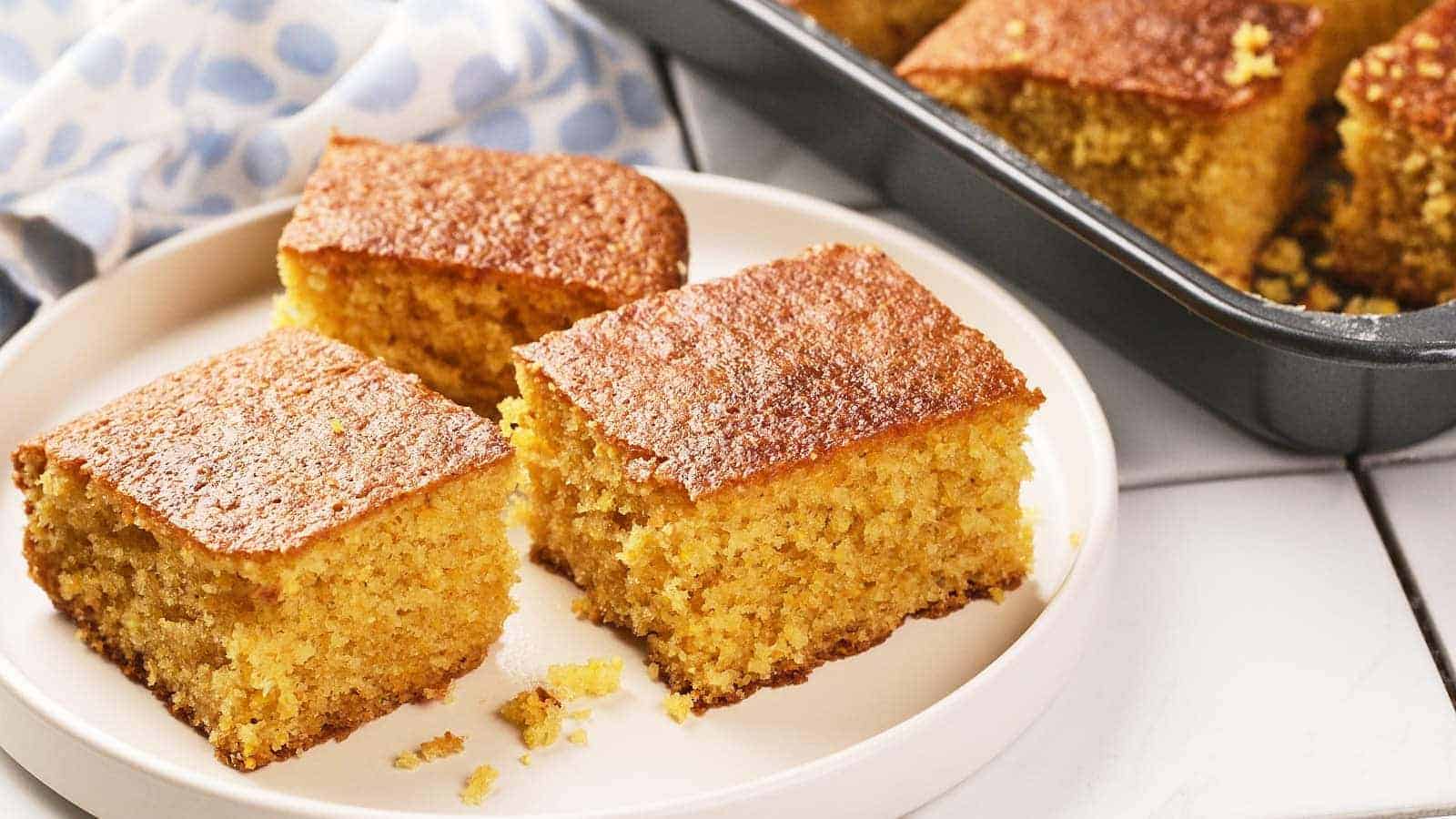 Three square pieces of cornbread are on a white plate, with more pieces visible in a baking pan in the background.