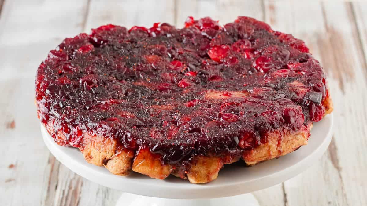 Cinnamon rolls with a caramelized cranberry topping sits on a white cake stand against a rustic wooden background.