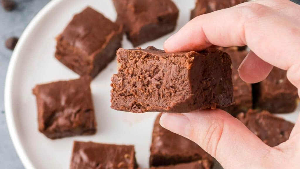 A hand holding a chocolate fudge with more brownies on a white plate in the background.