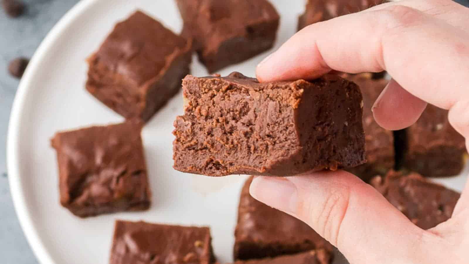 A hand holds a piece of chocolate fudge with a bite taken out, with more pieces of fudge on a white plate in the background.