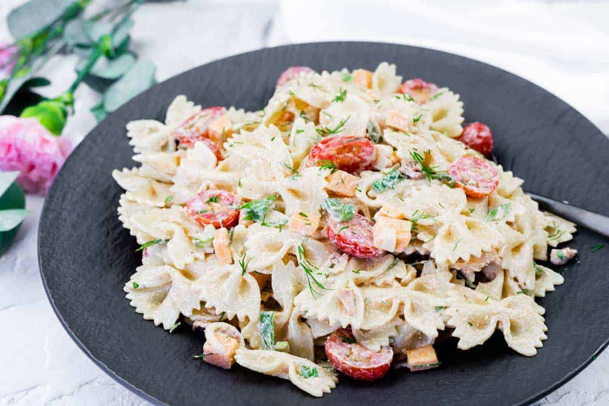 A black plate with bow-tie pasta salad mixed with cherry tomatoes, diced vegetables, herbs, and a creamy dressing.