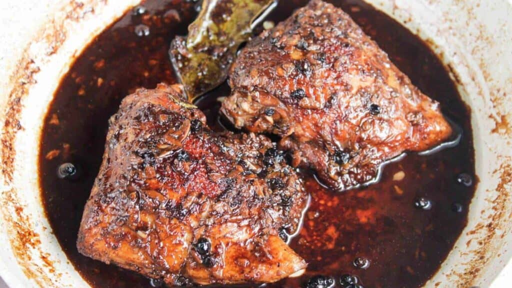 Two pieces of chicken simmer in a dark, seasoned sauce with visible peppercorns and a bay leaf in a pan.