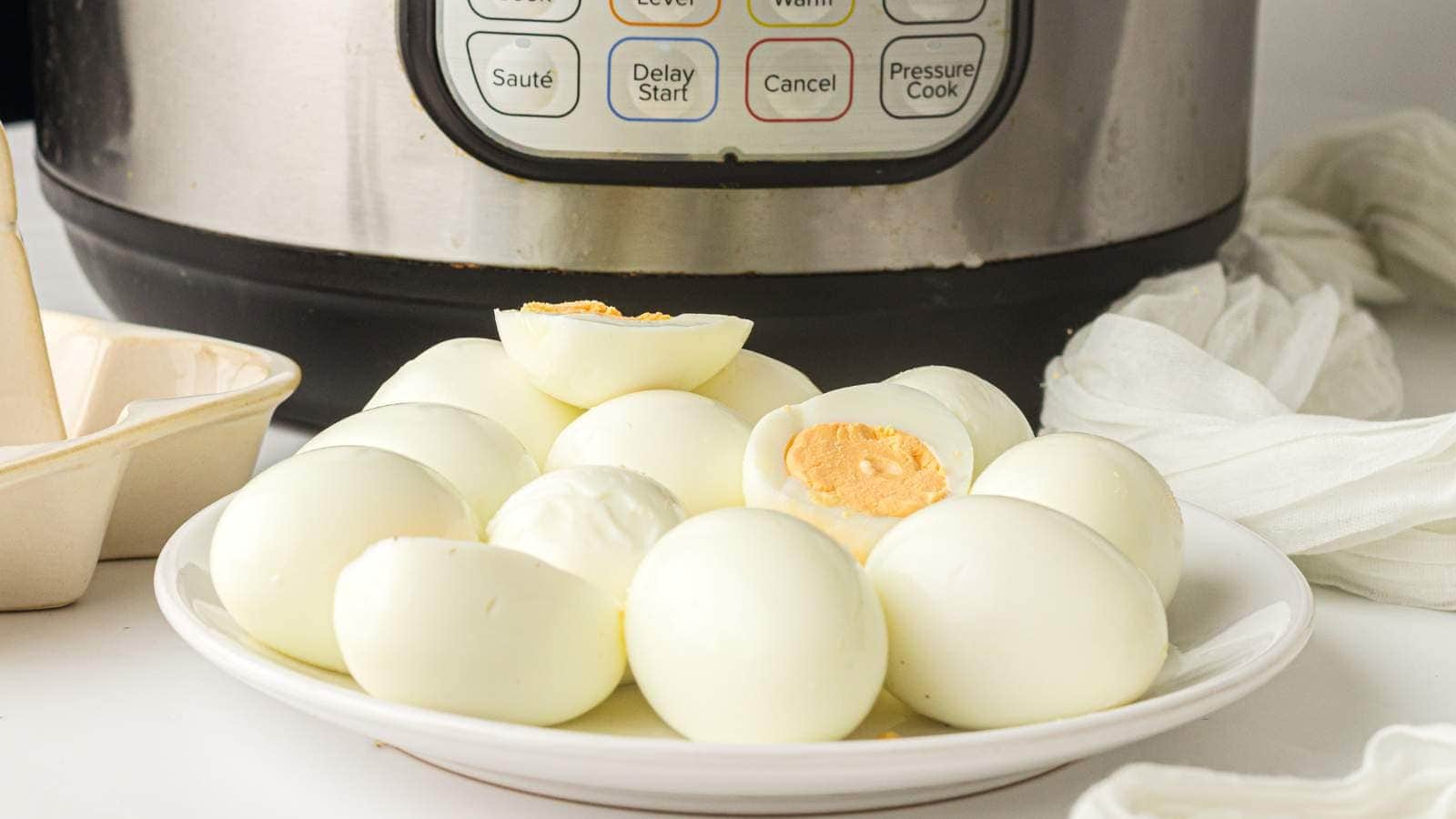 A plate of peeled hard-boiled eggs is placed in front of an instant pot on a white countertop.