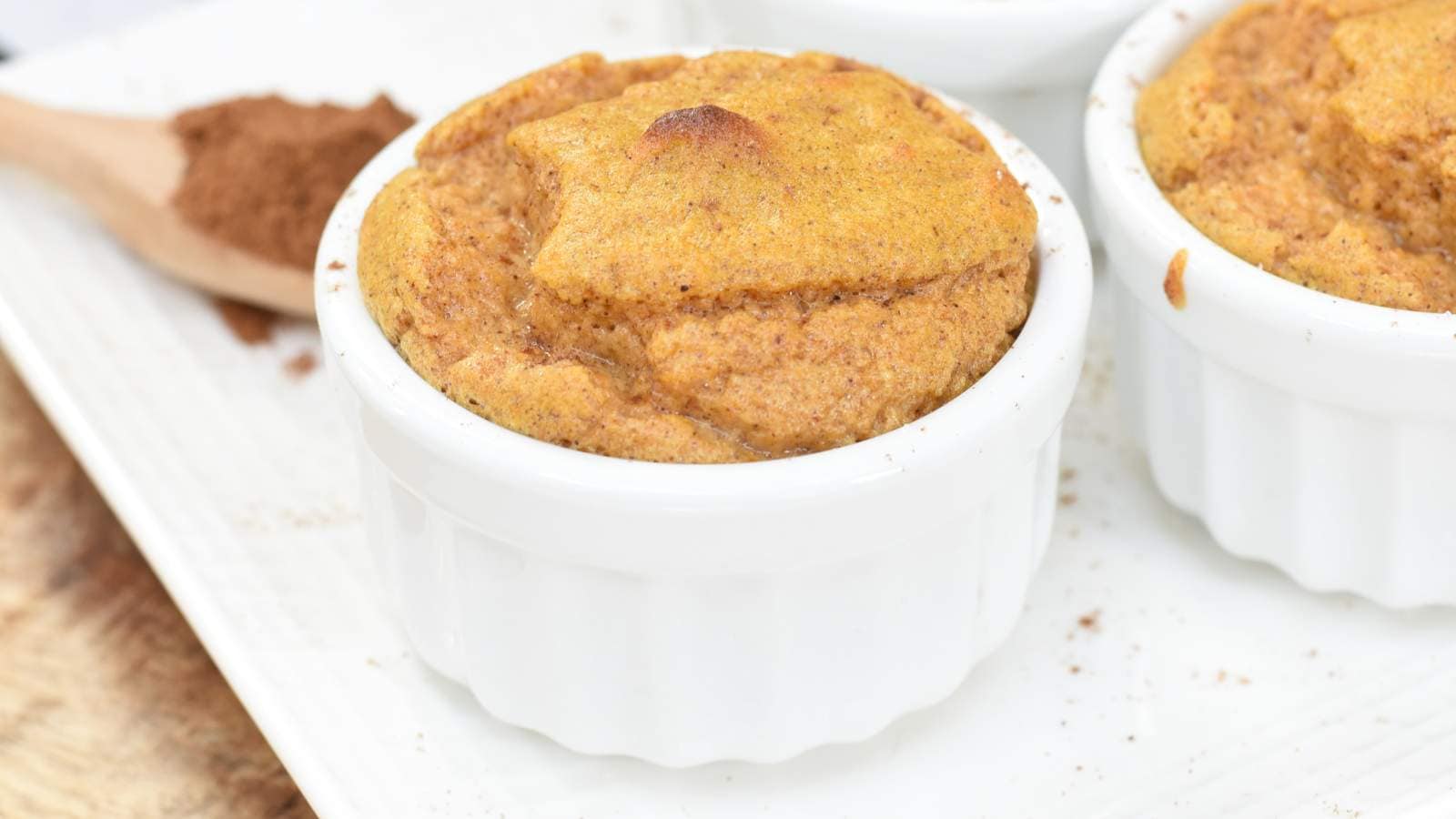A baked souffl&eacute; in a white ramekin sits on a white plate with a wooden spoon and ground cinnamon in the background.