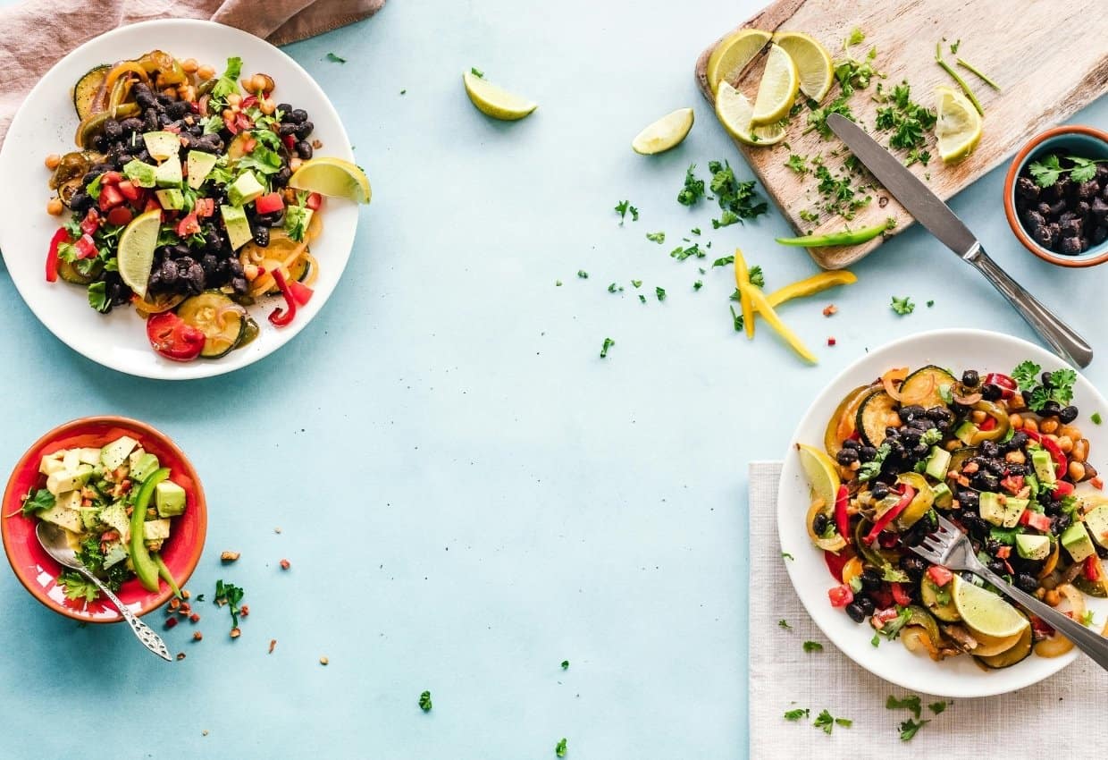Three plates of colorful salad with vegetables, black beans, avocado, and lime wedges are arranged on a light blue surface next to a chopping board, knife, herbs, and scattered lime slices.