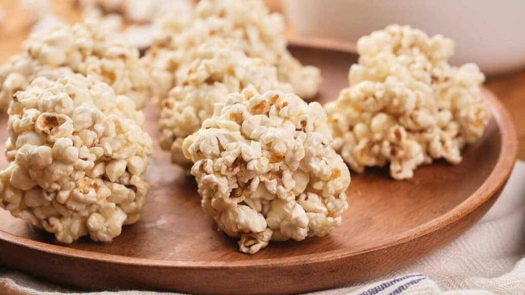 Six popcorn balls rest on a wooden plate, with a blurred background and part of a striped cloth visible underneath.