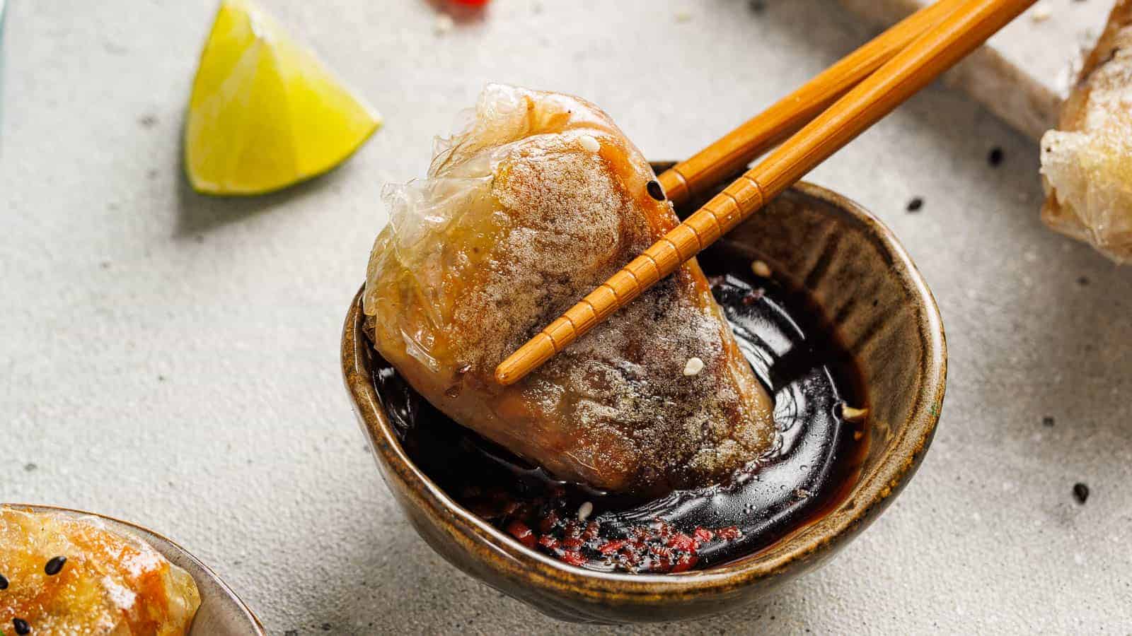 A pair of chopsticks holding a dumpling dipped in soy sauce, with a slice of lime and sesame seeds in the background.