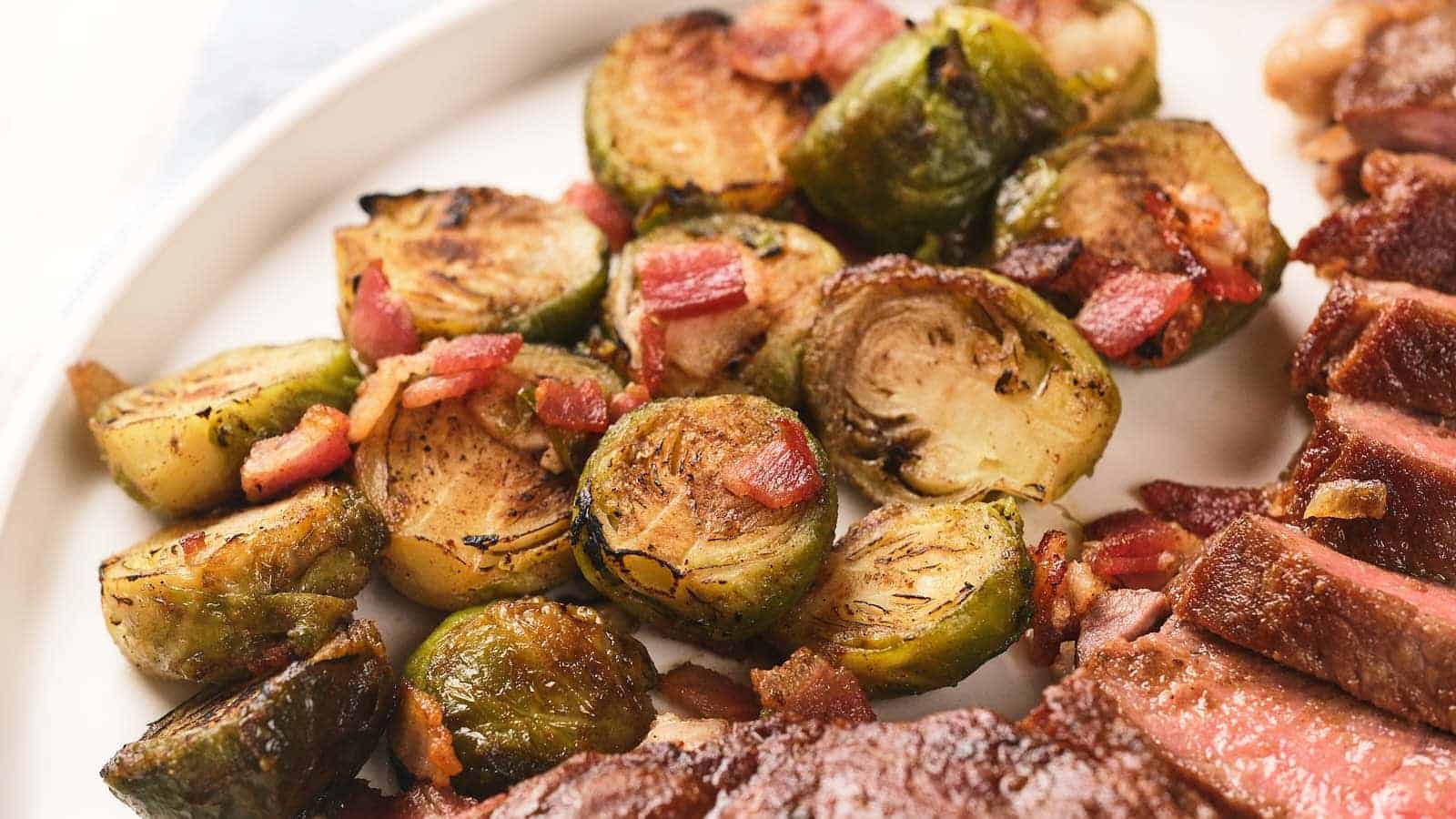 Close-up of roasted Brussels sprouts with bacon pieces on a white plate, next to slices of cooked meat.
