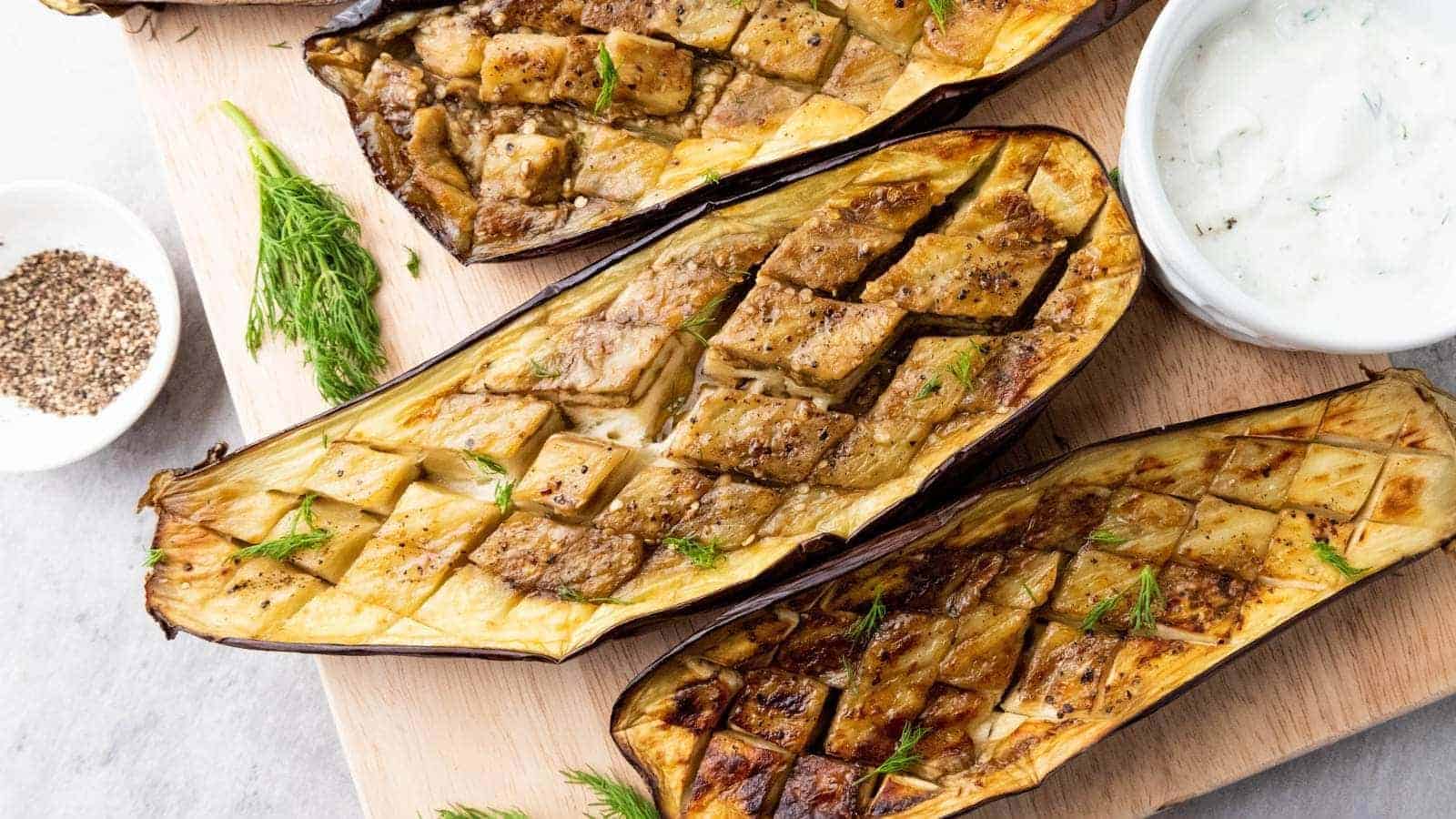 Three roasted eggplant halves scored in a crisscross pattern, garnished with herbs, served on a wooden board.
