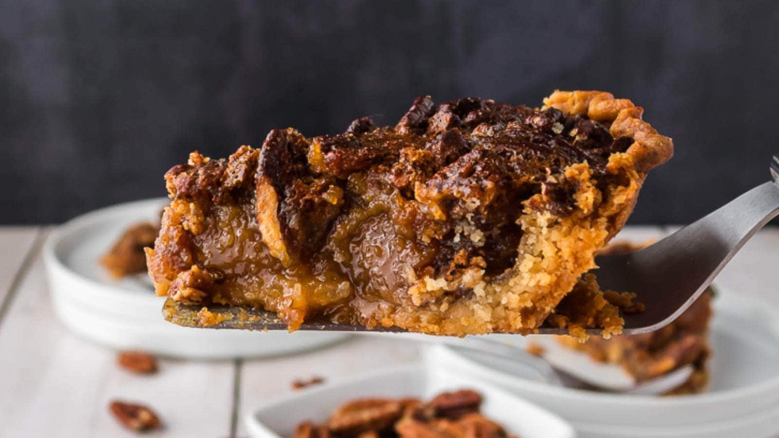 A close-up of a slice of pecan pie on a spatula, with a golden-brown crust and a gooey filling, held above a stack of white plates.