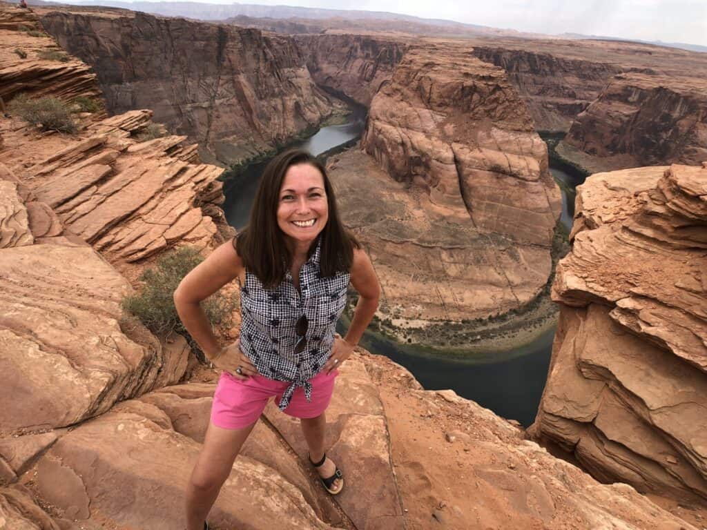 A woman stands and smiles on a rocky ledge overlooking Horseshoe Bend, where the Colorado River curves around a large rock formation in a desert landscape.