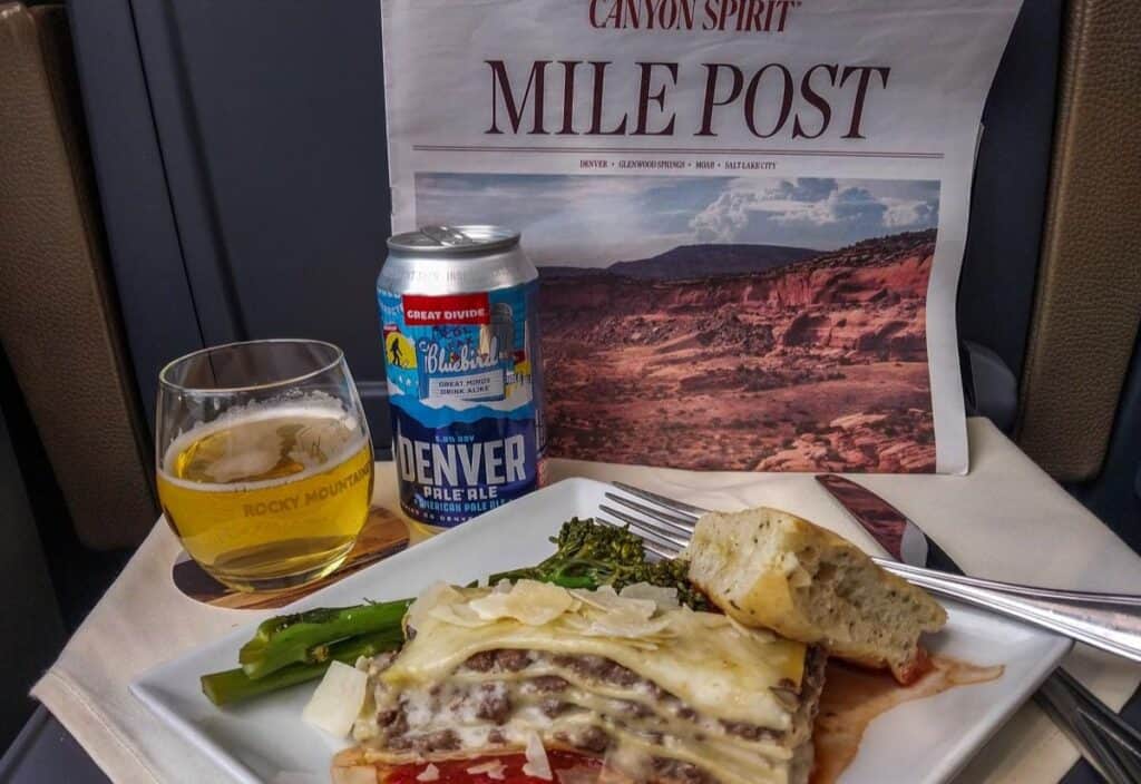 A meal of lasagna, broccolini, and bread is served with a glass of beer and a can of Denver Pale Ale, alongside a "Mile Post" newspaper featuring a canyon landscape that captures the true Canyon Spirit.