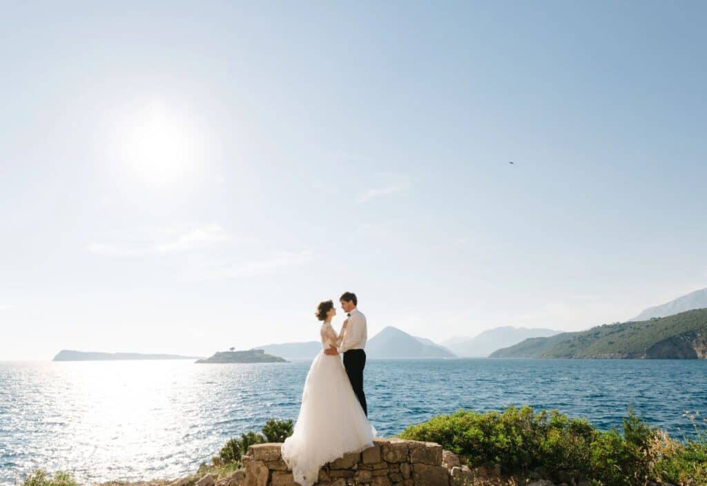 A bride and groom stand together on a stone ledge by the sea, with mountains, islands, and the sun in the background.