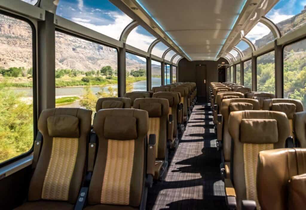 The interior of a Canyon Spirit train car with large panoramic windows, empty tan seats, and scenic countryside visible outside.