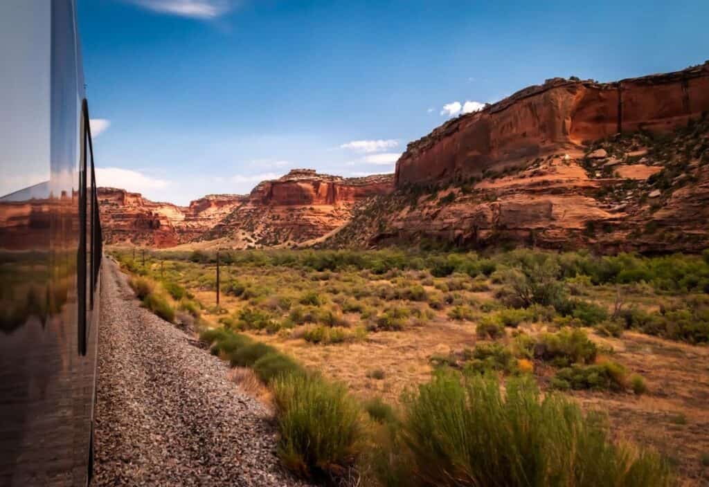 A train travels along tracks through a dry, rocky canyon landscape with red cliffs under a blue sky, capturing the true Canyon Spirit.