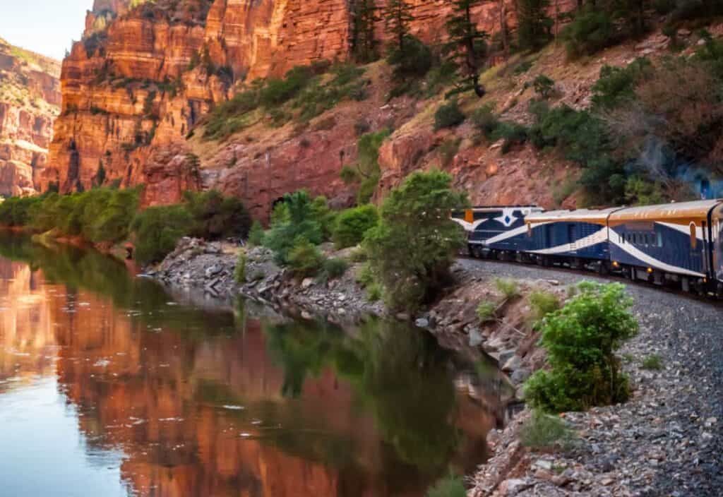 A passenger train travels along tracks beside a river, with steep red rock cliffs and green trees reflected in the water, capturing the essence of the Canyon Spirit.