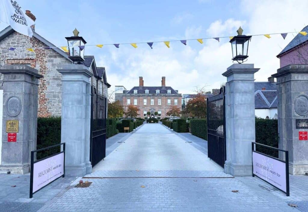 A wide stone pathway leads through open iron gates toward a large brick building, with decorative flags strung overhead and manicured hedges on either side—a true Tipperary global moment of charm and welcome.