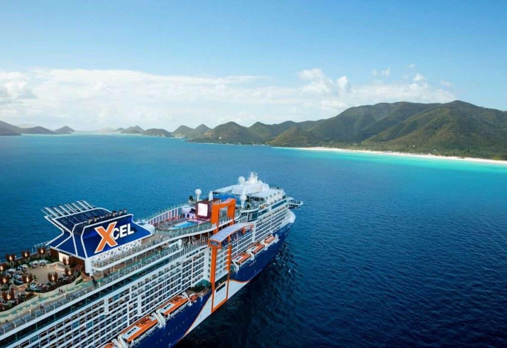 A large cruise ship labeled &ldquo;Xcel&rdquo; sails near a tropical coastline with green mountains and a sandy beach under a clear blue sky.
