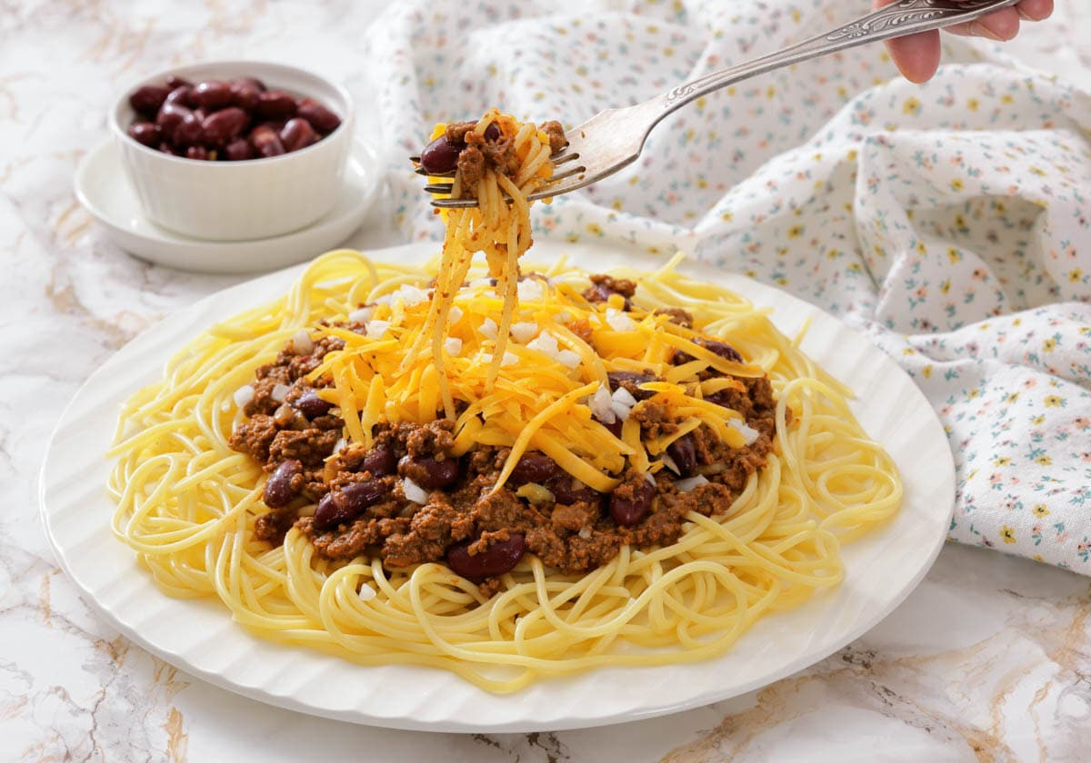 A plate of spaghetti topped with Cincinnati chili, shredded cheese, and beans, with a fork lifting a portion.
