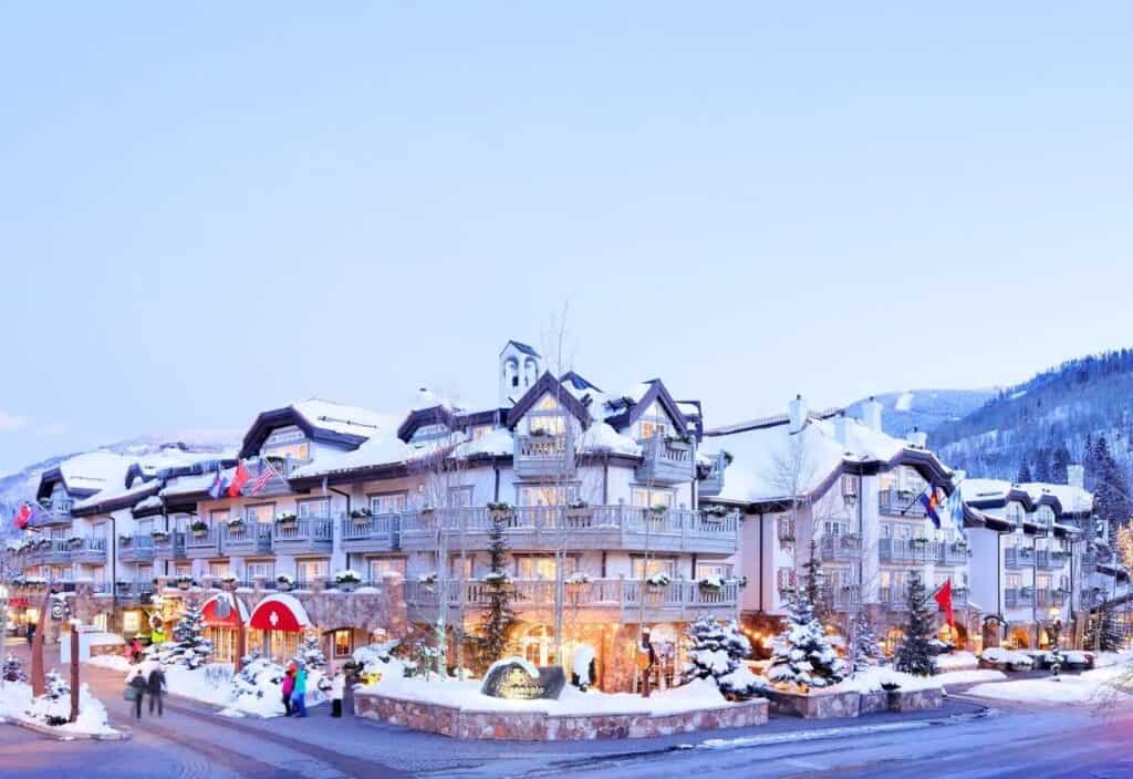 A large alpine-style hotel with balconies and snow-covered roofs, surrounded by snowy trees and mountains under a clear sky, stands as a new marker of quality. People can be seen near the entrance, adding life to this picturesque retreat.