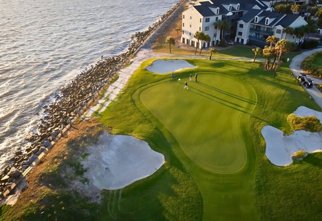 Aerial view of a coastal golf course green on Fripp Island with three golfers, sand bunkers, and nearby residential buildings next to the ocean.
