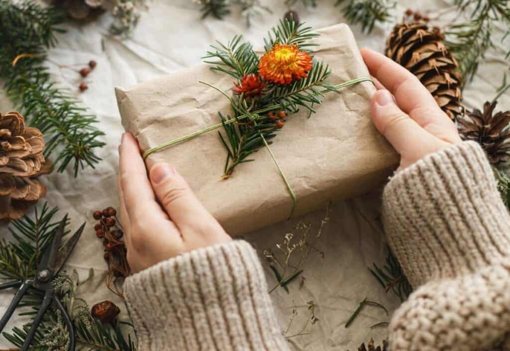 Hands holding a gift wrapped in brown paper and decorated with a yellow flower, pine sprig, and twine. Pine cones and greenery are scattered on the table.