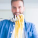 A person holding cooked spaghetti with a pasta fork, with the noodles in focus and the person blurred in the background.