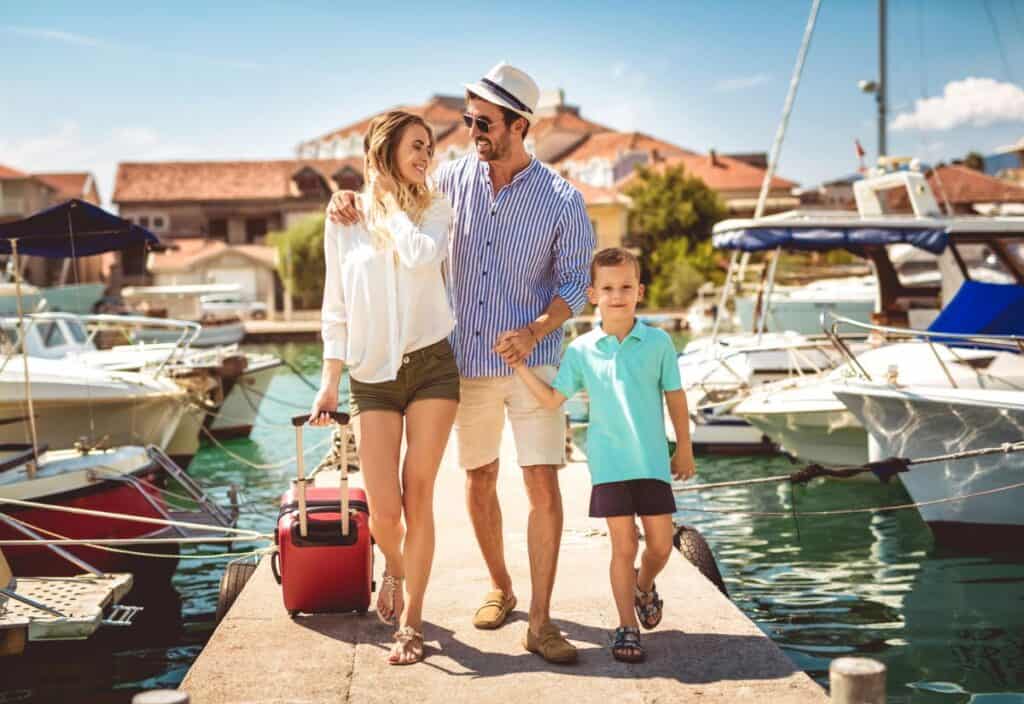 A family of three walks on a marina dock with boats in the background, embodying affordable high-end travel trends. The woman carries a suitcase, and all appear dressed for warm weather.