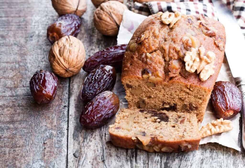 A loaf of date and walnut bread sits on a wooden surface, partially sliced, surrounded by whole dates and walnuts&mdash;perfect for celebrating National Date Nut Bread Day.