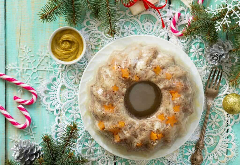 A festive aspic dish garnished with carrot stars sits on a white plate, surrounded by Christmas decorations, pine branches, and a bowl of mustard.