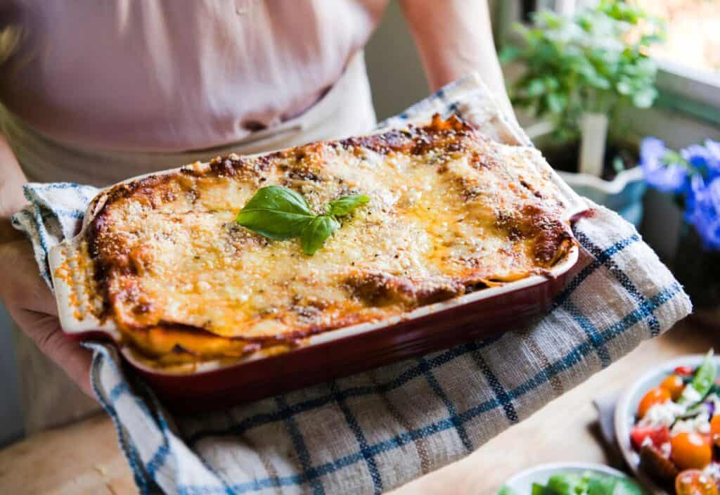 A person holds a freshly baked lasagna in a red dish, topped with basil, on a checkered kitchen towel near a window with plants.