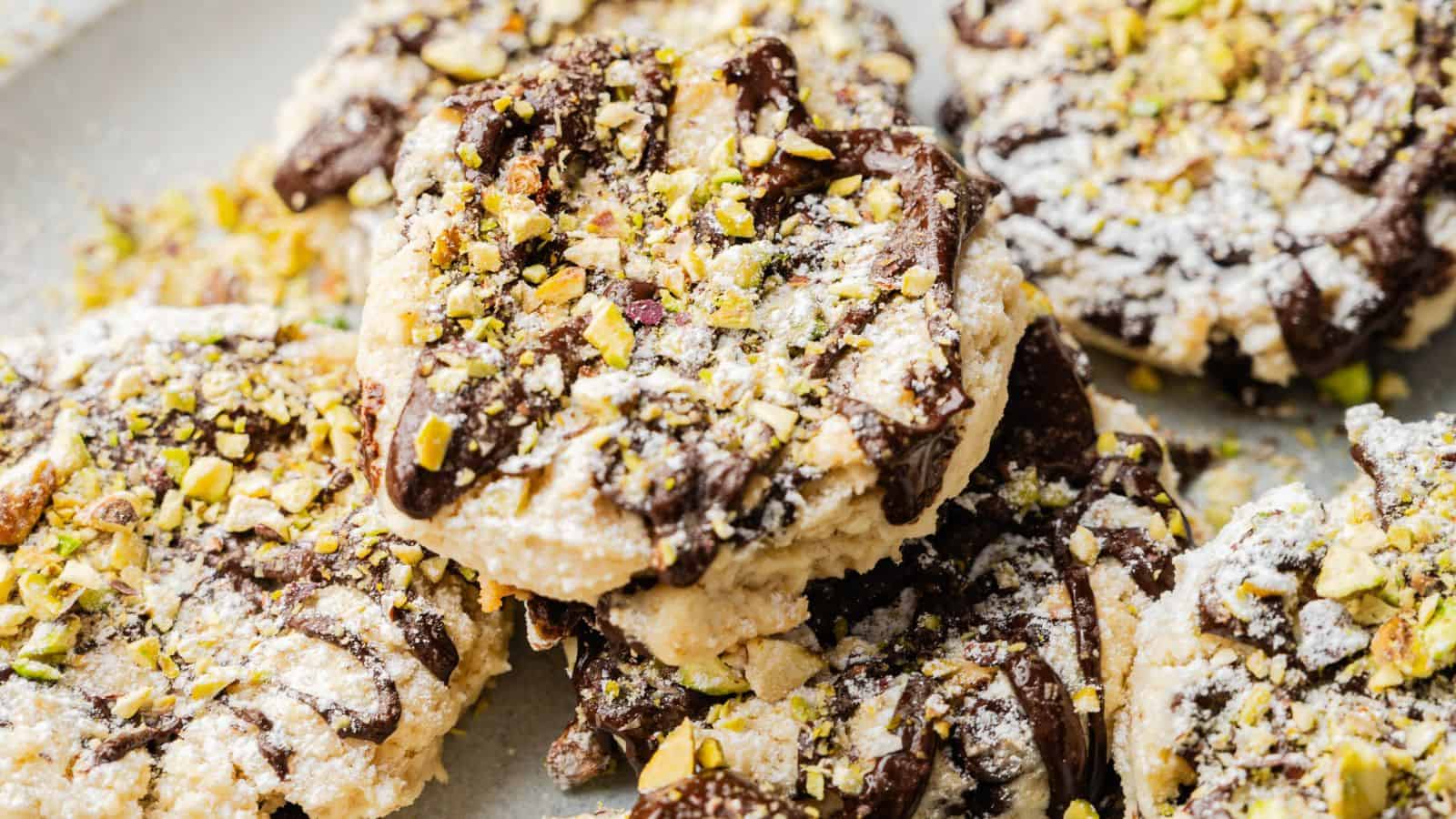 A close-up of cookies topped with powdered sugar, chopped pistachios, and drizzled with chocolate.