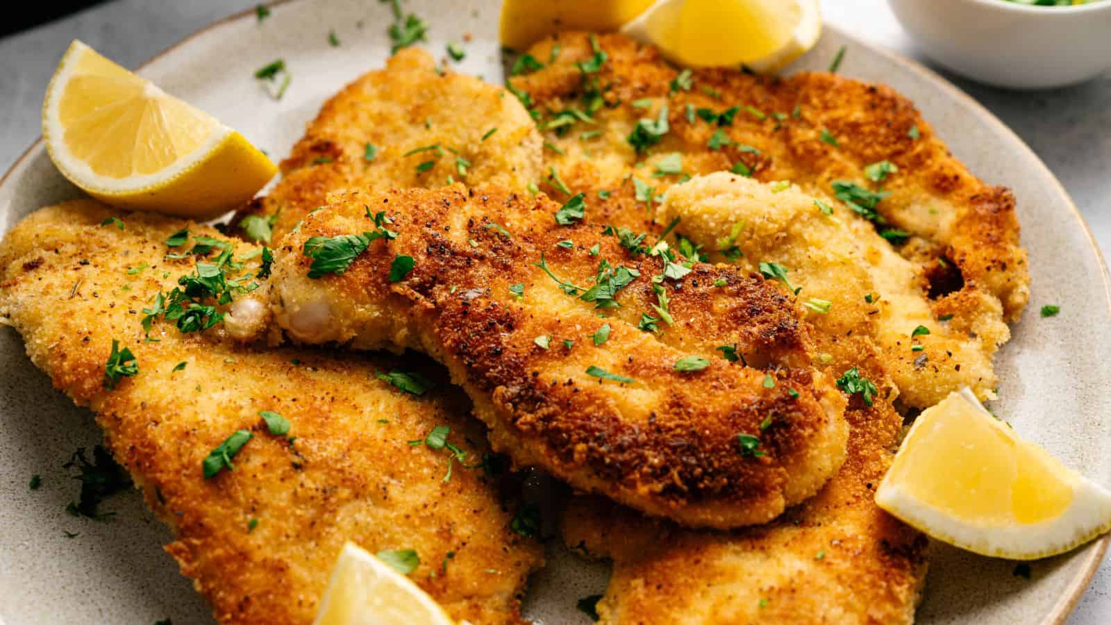 Plate of breaded and fried chicken cutlets garnished with chopped parsley and served with lemon wedges.